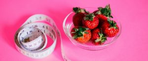 a measuring tape next to a glass bowl of strawberries with a pink background