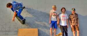 skateboarders chilling against a wall whilst one of them is doing skateboard tricks