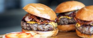 three meat, bacon and cheese burgers on a wooden cutting board
