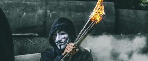 a man wearing an anonymous mask whilst holding a bundle of sticks on fire