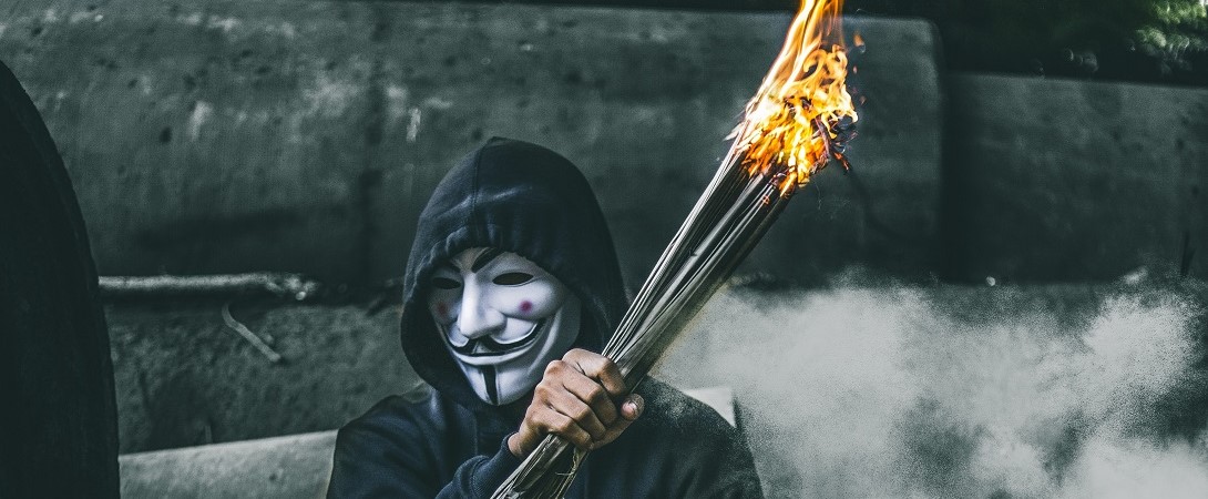a man wearing an anonymous mask whilst holding a bundle of sticks on fire