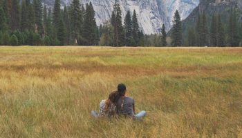 two woman sitting in a field in front of a forest and mountain range