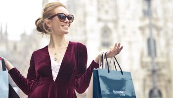 a blonde woman with glasses and a red dress walking around with several different sized and coloured shopping bags
