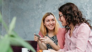 two women drinking coffee in a cafe on their phones and talking to each other