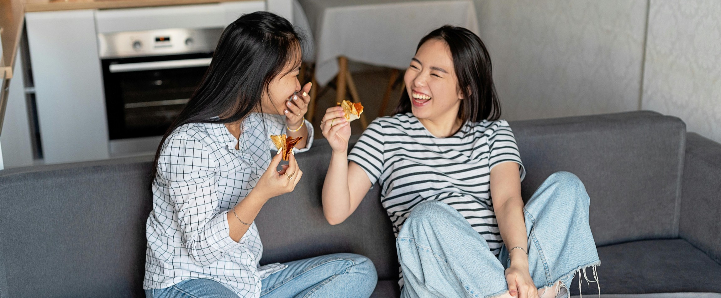 two women laughing eating pizza together on couch
